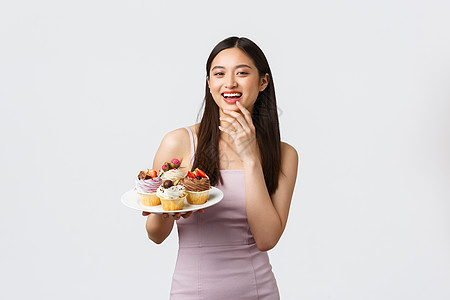 生活方式 假期 庆祝活动和食品概念 身着晚礼服的美丽亚洲年轻女性主持派对 用美味的甜点招待客人 拿着带纸杯蛋糕的盘子 白色背景图片