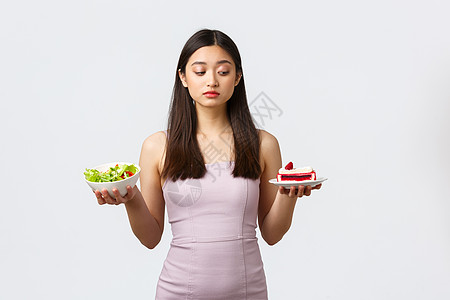 健康的生活方式 休闲和食品理念 美丽的亚洲女孩试图抵制诱惑 咬住充满卡路里的甜美可口蛋糕 坚持节食 吃沙拉 白色背景图片