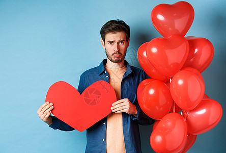 悲哀的人看着心碎和孤独 抱着纸红色的心脏 站在气球旁边 在蓝背景上站立 蓝色的 可爱的图片