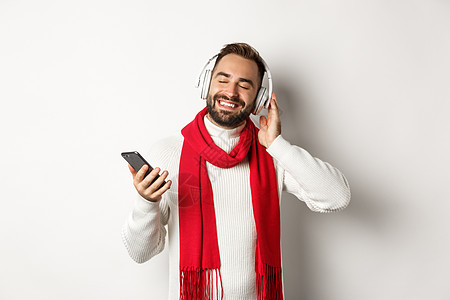 寒假和技术概念 满意的男人戴着耳机听音乐 闭着眼睛 高兴地微笑 拿着智能手机 白色背景 派对 庆典图片