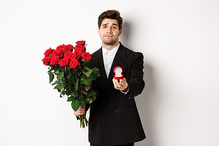 身着黑色西装 手持玫瑰和订婚戒指 站在白色背景下向他求婚的英俊微笑男子 浪漫图片