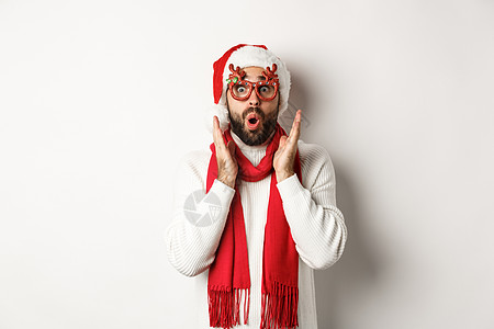 圣诞节 新年和庆祝活动的概念 戴着派对眼镜和圣诞老人帽子的男人看起来很惊讶 听到购物优惠 站在白色背景上 黑发 广告图片