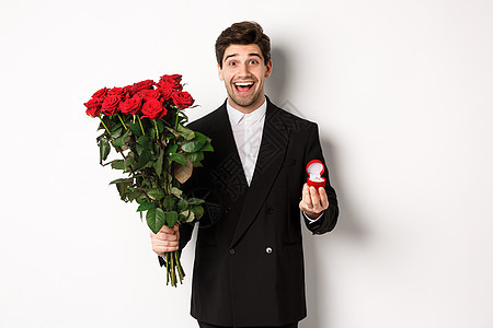 身着黑色西装 手持玫瑰和订婚戒指 站在白色背景下向他求婚的英俊微笑男子 爱 惊喜图片