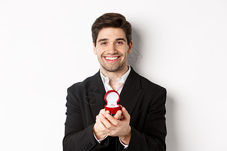 英俊男人看着浪漫的画面 用订婚戒指打开小盒子 提出一个建议和微笑 站在白色背景下站立 情人节 假期图片