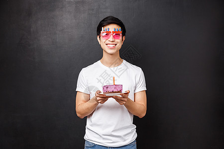 生日 庆典和派对概念 身穿白色 T 恤 快乐 快乐的亚洲年轻男子 戴着滑稽的眼镜 拿着带蜡烛的生日蛋糕 吹它许愿 微笑着兴奋 站图片