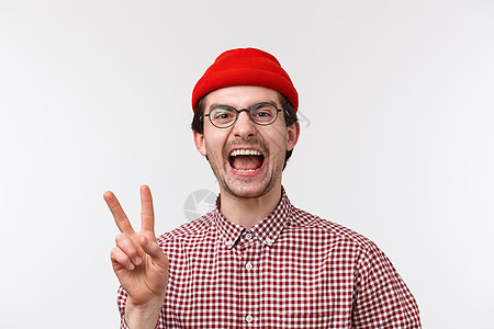 特写肖像快乐而有趣的白人留着胡须的男性戴着眼镜和红色的帽子 笑着展示和平的姿态 看起来相机很开心 站在白色背景图片