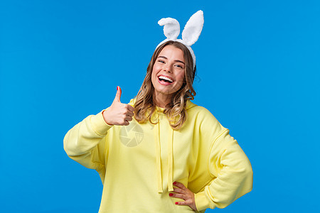 假期 传统和庆祝概念 乐观可爱的金发女孩 长着兔子耳朵 笑着摆出赞同的姿势 竖起大拇指称赞干得好 复活节快乐 蓝色背景图片
