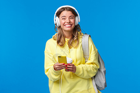 快乐漂亮的年轻女学生背着背包 戴着耳机听最喜欢的播放列表 无忧无虑地对着镜头笑笑 拿着手机 站在蓝色背景 喝 美丽的图片