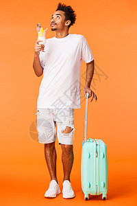 身穿白色 T 恤 站在行李旁 抬头看着太阳 喝鸡尾酒享受完美假期 站立橙色背景的全身垂直镜头梦幻 轻松的非洲裔美国人图片