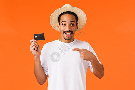 开朗快乐 面带微笑的非洲裔美国人在令人敬畏的新银行开设账户 指着信用卡 开心地笑着 就像客户服务一样 站立橙色背景图片