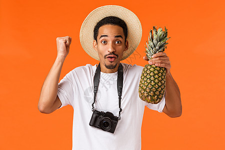 万岁假期和热带水果 欢快兴奋的非洲裔美国人终于出国旅行 在旅行中玩得开心 握着拳头 拿着菠萝 在橙色背景下拿着相机站着图片