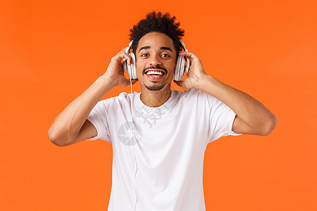 幸福 技术和小工具的概念 身穿白色 T 恤 戴着耳机听音乐 面带微笑的相机快乐 喜欢礼物 橙色背景的迷人快乐 魅力超凡的非洲裔美图片