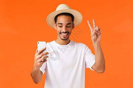 快乐可爱的非洲-美国度假男子自拍上网张贴 持有智能手机 在移动相机上微笑和做和平标志 站立橙色背景喜悦 令人赞叹的节日图片