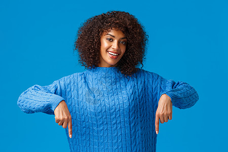 穿着冬季毛衣的可爱 友善和亲情的年轻非洲-美国女性 指向低点 邀请浏览网路购物绝佳网站 表示底广告 蓝背景图片
