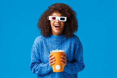 欢快华丽的非洲裔美国女性喜欢在电影院看电影 带着 3d 效果兴奋地笑笑 戴着眼镜 拿着爆米花 站在蓝色背景图片