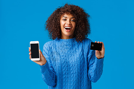 快乐而兴奋 迷人的非洲裔美国女性购物狂在网上购买礼物 展示申请 购物应用程序和信用卡 笑着笑着 站在蓝色背景图片