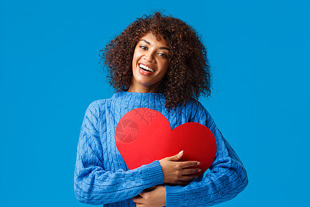 可爱而温柔风趣 面带微笑 留着爆炸式发型的非洲裔美国女性 将大红色心形标志按在胸前 用愉悦迷人的笑容拥抱它 表现出爱和感情 蓝色背景