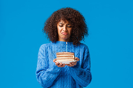 心烦意乱 忧郁 沮丧的年轻非洲裔美国女性讨厌庆祝生日 感觉自己变老了 看着生日蛋糕上点着蜡烛 生闷气 蓝色背景 看起来很烦恼和不图片