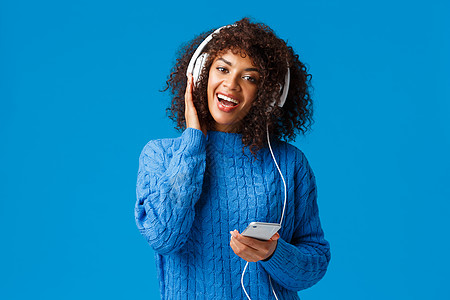 快乐开朗的非洲裔美国女性在智能手机上玩卡拉 OK 游戏应用程序 戴着耳机唱歌 享受美妙的音乐 音质 站立的蓝色背景图片