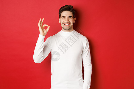 冬天假期 圣诞节和生活方式的概念 穿着白色毛衣的英俊男子快乐 他们展示了良好的标志 微笑喜悦 推荐商店 站在红色背景之上 购物 图片