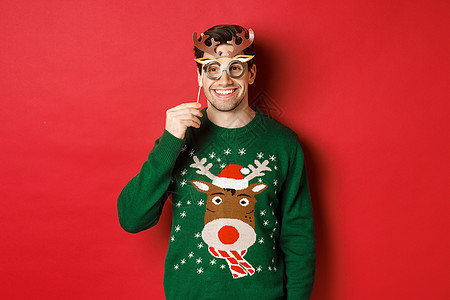 穿着圣诞毛衣的帅帅帅帅男人 戴着党面罩微笑 享受新年庆典 站在红背景上 男生 购物图片