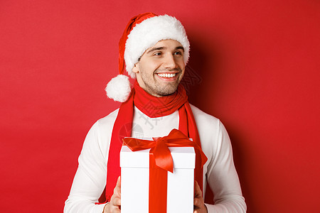 冬假 圣诞节和生活方式的概念 穿着圣塔帽和围巾的有魅力男子近亲 持有新年礼物 向右看并微笑 站在红色背景之上 成人 购物图片