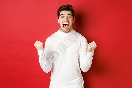 穿着白色毛衣的快乐帅哥的形象 赢得了一些东西 握拳 微笑着惊讶 庆祝胜利 站在红色背景上 黑发 成就图片