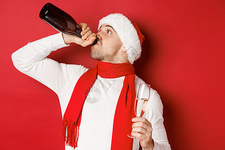 寒假 圣诞节和生活方式的概念 男人在新年派对上喝醉 喝着瓶子里的香槟 戴着圣诞帽 站在红色背景上 大胡子 美丽的图片