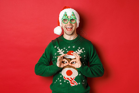 穿着圣塔帽和派对眼镜的无忧无虑英俊男子肖像 取笑他的圣诞节毛衣 在红色背景下看着快乐 庆典图片