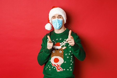 新年 covid-19 和社会疏远的概念 穿着圣诞毛衣 医用面具和圣诞帽的快乐男人 用手指指着镜头 祝节日快乐 站在红色背景上 图片