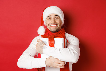 寒假 圣诞节和生活方式的概念 戴着圣诞帽和围巾的可爱男人的形象 拥抱他的新年礼物 微笑着受宠若惊 站在红色背景上 庆祝 圣诞节快背景图片