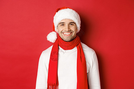 寒假 圣诞节和生活方式的概念 留着鬃毛的帅哥的特写 戴着带围巾的圣诞帽 笑得很开心 祝新年快乐 红色背景 微笑 黑发背景图片