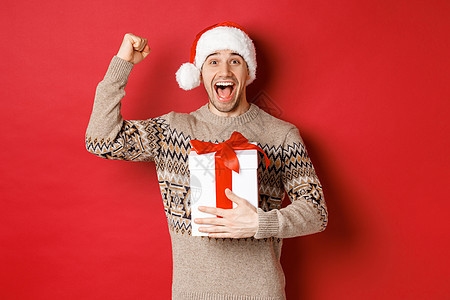 快乐而兴奋的帅哥的形象 reicing 圣诞礼物 胜利地举起手 微笑着 庆祝新年 站在红色背景上 戴着圣诞老人的帽子 成就 派对图片