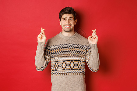 希望和担忧的穿着圣诞节毛衣的人等待着某事 交叉手指以换取好运和许愿 紧张于新年礼物 红色背景 笑声 成人 工作室背景图片