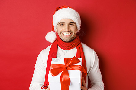 寒假 圣诞节和生活方式的概念 戴着圣诞帽和围巾 拿着礼物 送新年礼物和微笑 红色背景的留着胡须的帅哥的特写 工作室 圣诞节快乐背景图片