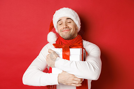 寒假 圣诞节和生活方式的概念 戴着圣诞帽和围巾的可爱男人的形象 拥抱他的新年礼物 微笑着受宠若惊 站在红色背景上 情感 成人背景图片