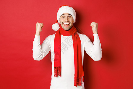 戴着圣诞帽和围巾的快乐而兴奋的男人的画像 欢欣鼓舞并赢得了一些东西 庆祝新年 站在红色背景上 黑发 冬天图片