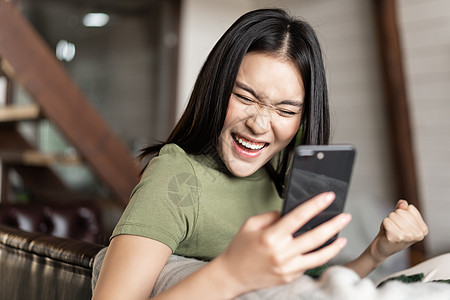 微笑的年轻女性看着满意 做拳手泵和阅读手机 坐在家里并取得胜利 休息 放松图片