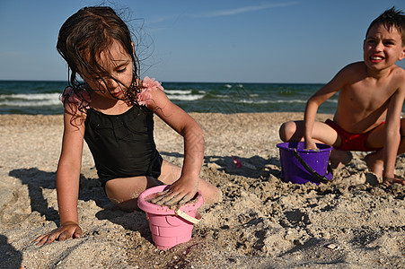 可爱的女孩用沙子填满了粉红色塑料玩具桶 以创造雕像和建造一个沙城堡 同时在海洋背景下与她的兄弟一起玩耍图片