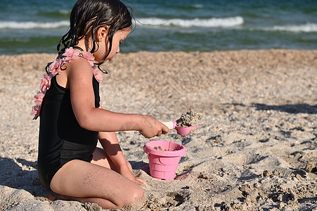 学龄前女孩的侧面肖像 她拿着一只子 用沙子填满粉红色玩具桶 建造沙质形状和城堡 享受日光浴和暑假图片