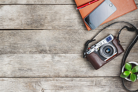 带有棕色组织者红笔的经典相机 放在一张灰色的木制复古桌子上 桌子上有电话和绿色植物 旅行中摄影师列表的概念 乡村风格的自由空间 图片