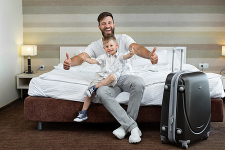 一个年幼的父亲和儿子坐在旅馆的床上 带着一个手提箱大笑图片