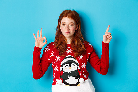圣诞快乐 穿着Xmas毛衣的红头红发女孩 指向右上角 展示新年宣战和赞成 赞美产品 圣诞节 脸图片