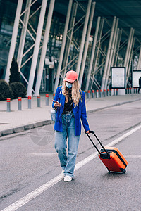 一名戴面罩 带着橙色手提箱在机场附近行走的女旅行者肖像 身穿蓝色牛仔裤和外套的年轻时尚女子 金发 套装图片