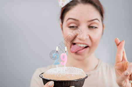 欢乐的caucasian女人露出舌头 用手指交叉在蛋糕上吹蜡烛 女孩庆祝她29岁生日 29日 漂亮的图片