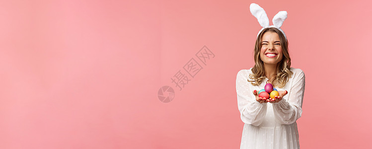 假期 春天和聚会的概念 兴奋迷人的金发女人的肖像 带着可爱的快乐微笑 拿着彩绘鸡蛋 穿着复活节服装 兔耳和白色连衣裙 粉红色背景图片