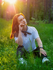 一位年轻女子在日落的背景下 在夏天坐在绿草的青草上坐落在森林中图片