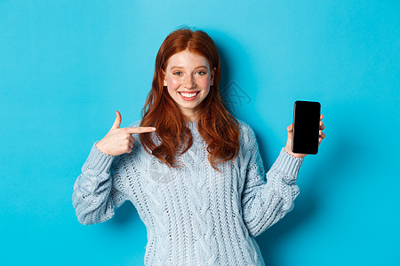 满意的红发女孩指着电话屏幕 显示智能手机应用程序或在线宣传和微笑 穿着蓝底毛衣站立在蓝色背景上 时尚 美丽图片