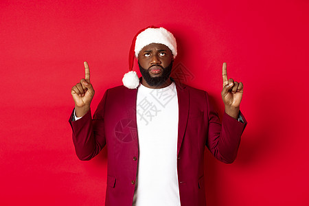 圣诞节 聚会和假期的概念 戴着圣诞帽的非洲裔美国人心烦意乱地抱怨 指着手指 失望地皱着眉头 站在红色背景下 购物图片
