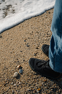 穿着黑鞋和蓝色牛仔裤的人的腿 在日落时海浪笼罩沙滩上站立图片